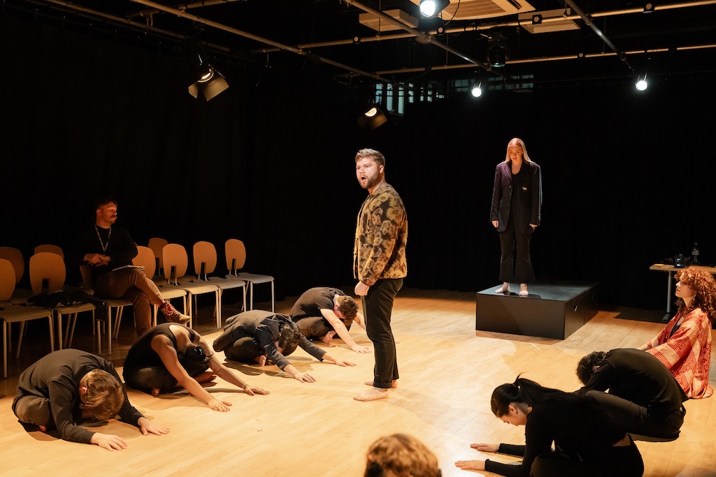 An actor wearing a patterned fur coat stands between two groups of actors who are sitting or kneeling on either side.